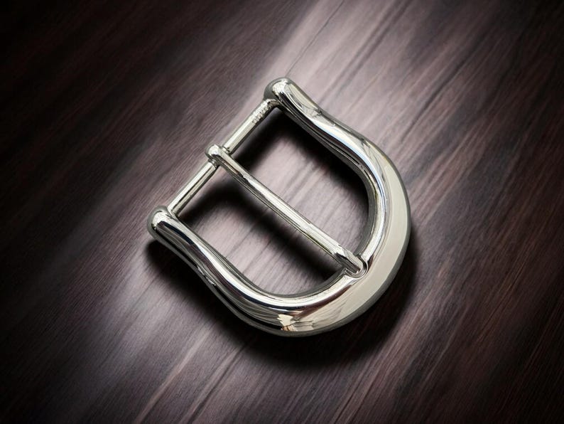 May include: A silver metal belt buckle with a smooth, polished finish. The buckle has a simple, classic design with a rounded shape.