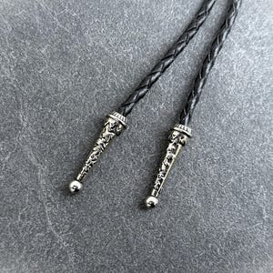 May include: Close-up of a bolo tie with a braided black leather cord. The silver-colored tips are ornate with a detailed design and a small, round ball at the end. The background is a textured gray surface.