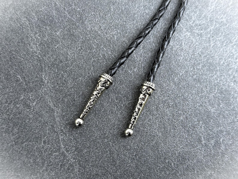 May include: Close-up of a bolo tie with a braided black leather cord. The silver-colored tips are ornate with a detailed design and a small, round ball at the end. The background is a textured gray surface.