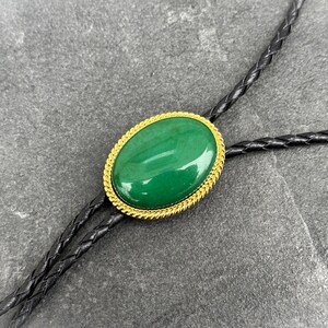 May include: Oval-shaped green stone bolo tie with a gold-colored rope-like border. The stone is set against a black braided leather cord. The bolo tie is a classic accessory.