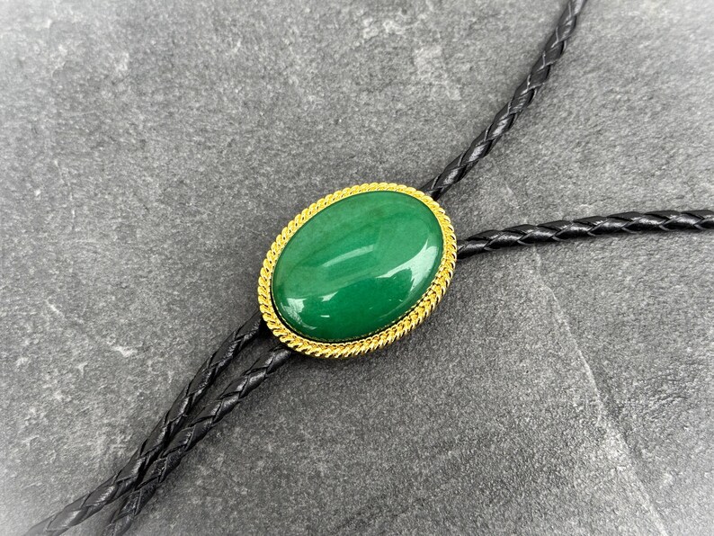 May include: Oval-shaped green stone bolo tie with a gold-colored rope-like border. The stone is set against a black braided leather cord. The bolo tie is a classic accessory.
