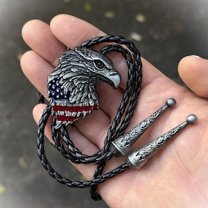 May include: A silver-colored bolo tie featuring an eagle head with the American flag design. The eagle is detailed with textured feathers and a red, white, and blue flag. The black braided cord has decorative silver tips.