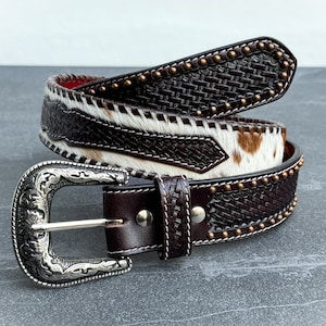 May include: A brown leather belt with a woven pattern and a silver buckle. The belt is decorated with brass studs and has a white and brown cowhide pattern.