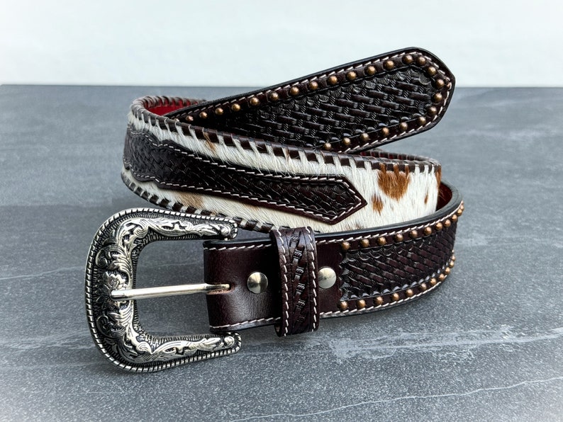 May include: A brown leather belt with a woven pattern and a silver buckle. The belt is decorated with brass studs and has a white and brown cowhide pattern.