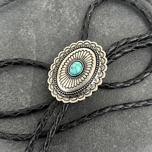 May include: A silver-toned bolo tie with a turquoise stone center. The oval pendant has a decorative edge and radial design. The black braided cord is looped and rests on a gray surface. This western-style accessory is suitable for various outfits.