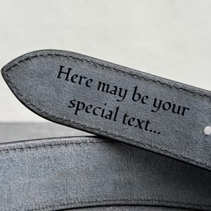 Op de afbeelding: Close-up van een grijze leren riem met de tekst "Here may be your special text..." gegraveerd op de punt. De riem heeft een enkel gat en een gestikte rand, wat duidt op een gepersonaliseerd accessoire.