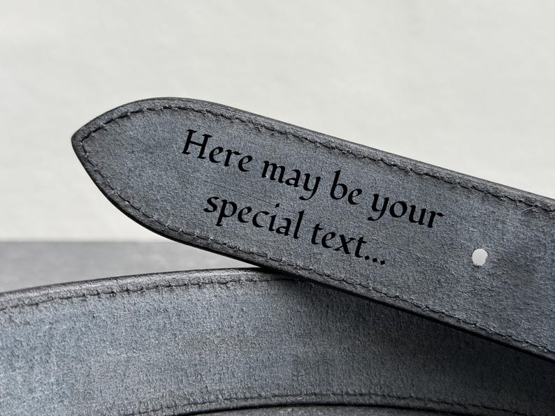 Op de afbeelding: Close-up van een grijze leren riem met de tekst "Here may be your special text..." gegraveerd op de punt. De riem heeft een enkel gat en een gestikte rand, wat duidt op een gepersonaliseerd accessoire.