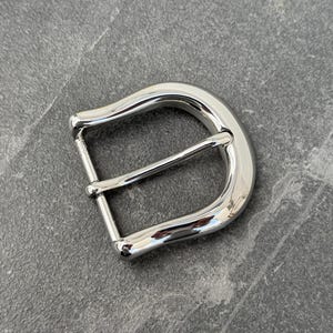 May include: A silver metal belt buckle with a smooth, rounded finish. The buckle has a single prong and a D-ring.