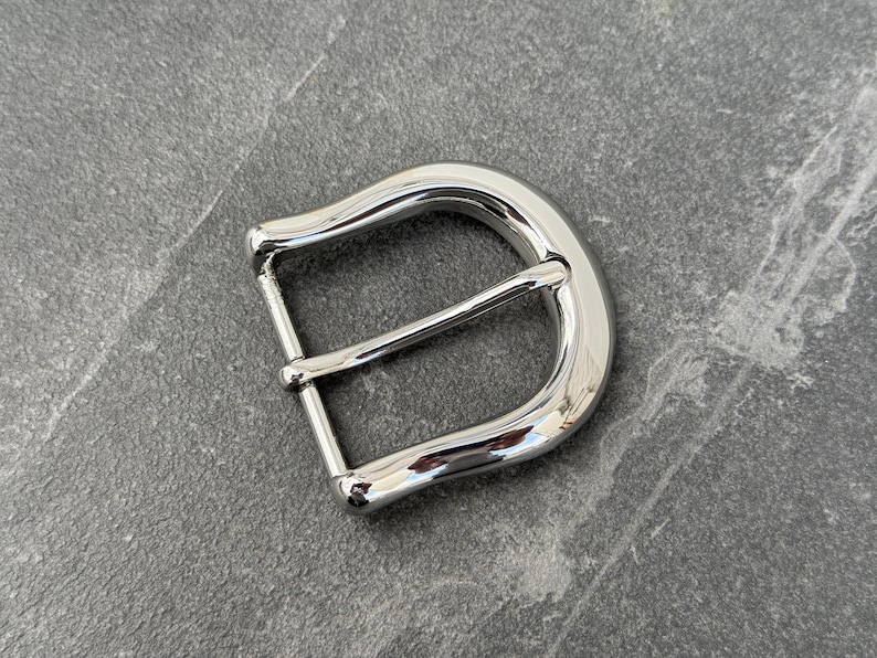 May include: A silver metal belt buckle with a smooth, rounded finish. The buckle has a single prong and a D-ring.