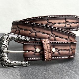 Op de afbeelding: Een bruine leren riem met een zilveren gesp. De riem heeft een prikkeldraadontwerp en witte stiksels. De gesp heeft een sierlijk bloemmotief. Een stijlvol accessoire voor dagelijks gebruik.