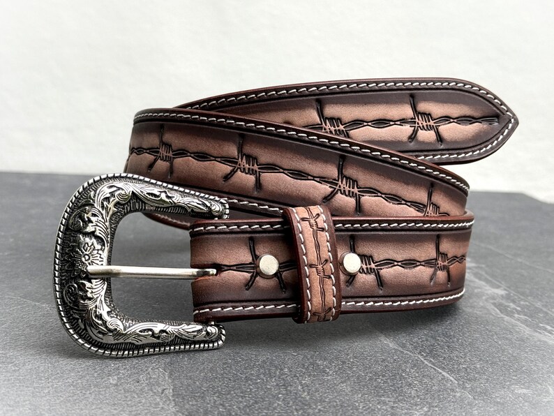 Op de afbeelding: Een bruine leren riem met een zilveren gesp. De riem heeft een prikkeldraadontwerp en witte stiksels. De gesp heeft een sierlijk bloemmotief. Een stijlvol accessoire voor dagelijks gebruik.