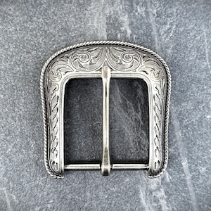 Puede incluir: Hebilla de cintur&oacute;n plateada con un dise&ntilde;o grabado y ornamentado. La hebilla presenta una abertura rectangular con una barra central y un borde decorativo. El patr&oacute;n detallado incluye motivos florales y de volutas, con un borde similar a una cuerda.