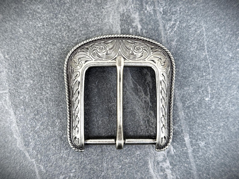 Puede incluir: Hebilla de cintur&oacute;n plateada con un dise&ntilde;o grabado y ornamentado. La hebilla presenta una abertura rectangular con una barra central y un borde decorativo. El patr&oacute;n detallado incluye motivos florales y de volutas, con un borde similar a una cuerda.