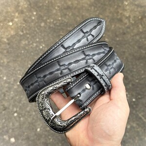 Op de afbeelding: Een zwarte leren riem met een zilveren gesp. De riem heeft een prikkeldraadontwerp en witte stiksels. De gesp heeft een sierlijk bloemmotief. De riem wordt in een hand gehouden.
