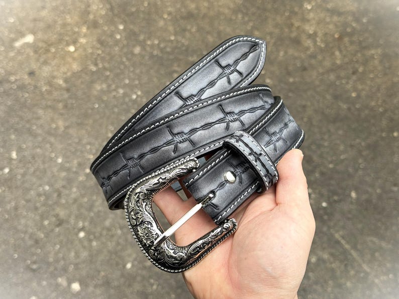 Op de afbeelding: Een zwarte leren riem met een zilveren gesp. De riem heeft een prikkeldraadontwerp en witte stiksels. De gesp heeft een sierlijk bloemmotief. De riem wordt in een hand gehouden.