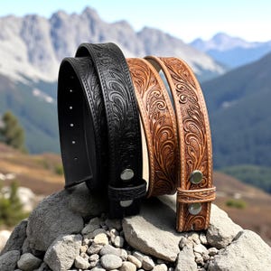 以下が含まれることがあります： フローラル柄のエンボス加工が施された3本の革製ベルト。1本は黒、他の2本は茶色です。ベルトは岩の表面に置かれ、背景には山が見えます。各ベルトにはシルバー色のバックルが付いています。