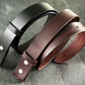 Puede incluir: Dos cinturones de cuero, uno negro y otro marrón, se muestran sobre una superficie gris. Los cinturones están hechos de cuero grueso y tienen remaches metálicos.