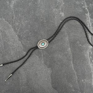 May include: A bolo tie with a silver-colored oval pendant featuring a turquoise-colored stone in the center. The pendant is surrounded by a decorative border. The black braided cord has silver-colored tips.