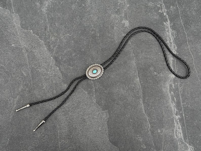 May include: A bolo tie with a silver-colored oval pendant featuring a turquoise-colored stone in the center. The pendant is surrounded by a decorative border. The black braided cord has silver-colored tips.