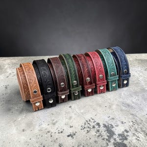 May include: A collection of colorful leather belts with floral tooling. The belts are in various colors, including brown, black, green, red, and blue. Each belt has a silver buckle and white stitching. The belts are arranged in a row, showcasing their different hues and designs.