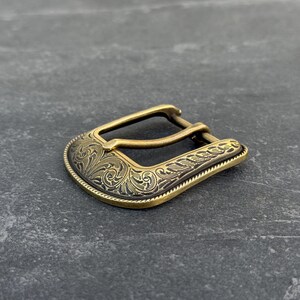 Puede incluir: Hebilla de cintur&oacute;n de color lat&oacute;n con un dise&ntilde;o grabado y ornamentado. La hebilla tiene una forma curvada con un detallado patr&oacute;n floral y un borde texturizado. El metal tiene un aspecto vintage y antiguo.
