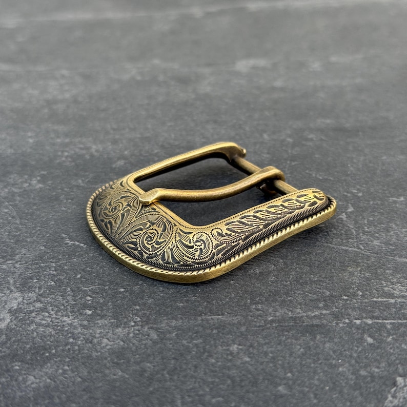 Puede incluir: Hebilla de cintur&oacute;n de color lat&oacute;n con un dise&ntilde;o grabado y ornamentado. La hebilla tiene una forma curvada con un detallado patr&oacute;n floral y un borde texturizado. El metal tiene un aspecto vintage y antiguo.
