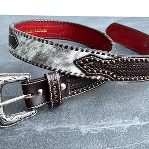 May include: A brown leather belt with a silver buckle and a white and brown patterned design. The belt has a row of gold studs along the edge.