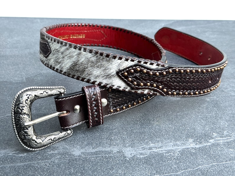 May include: A brown leather belt with a silver buckle and a white and brown patterned design. The belt has a row of gold studs along the edge.