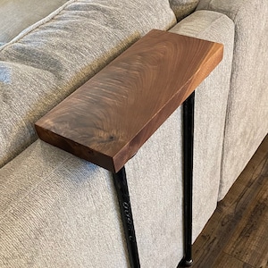 May include: A wooden side table with a dark brown walnut top and black metal legs. The table is attached to a gray couch.