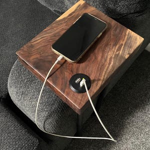 May include: A wooden side table with a phone charging on it. The table has a built-in charging port with two USB ports. The phone is connected to the charging port with a white cable.
