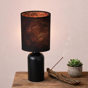 Può includere: Una lampada da tavolo nera con base cilindrica e paralume con un motivo scuro. La lampada è su una superficie di legno, accanto a un porta incenso in ottone e una piccola pianta succulenta in vaso.
