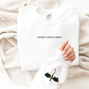 May include: White sweatshirt with the text "VICIOUS LITTLE THING" printed in black. The sleeve features a black and yellow dragon graphic. The sweatshirt is folded on a neutral-coloured surface.
