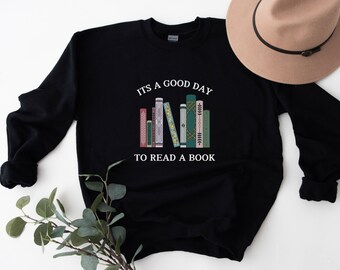 May include: Black sweatshirt with the text "IT'S A GOOD DAY TO READ A BOOK" above a graphic of colorful books. The sweatshirt is a crew neck style and has long sleeves. A tan hat is visible in the background.