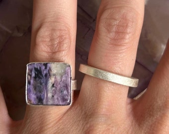 May include: A silver ring with a square purple and white stone set in the center. The ring is worn on the index finger of a hand.