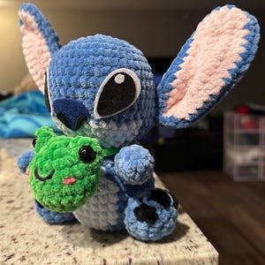 May include: A blue crocheted plush toy with large ears and black eyes, holding a green crocheted frog. The plush has a textured, fuzzy appearance. The frog has black eyes and a smiling mouth. The toy is sitting on a light-colored surface.