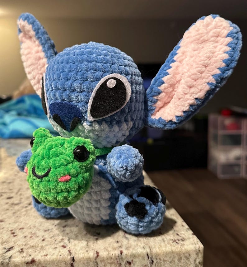 May include: A blue crocheted plush toy with large ears and black eyes, holding a green crocheted frog. The plush has a textured, fuzzy appearance. The frog has black eyes and a smiling mouth. The toy is sitting on a light-colored surface.