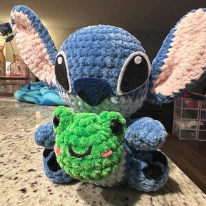 May include: A handmade blue crochet plush toy resembling a cartoon character, with large ears and black eyes. It's holding a green crochet frog with black eyes and a smiling face. The plush has pink accents and is made of soft yarn.