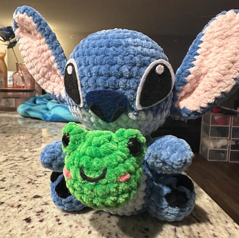 May include: A handmade blue crochet plush toy resembling a cartoon character, with large ears and black eyes. It's holding a green crochet frog with black eyes and a smiling face. The plush has pink accents and is made of soft yarn.