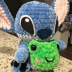 May include: A plush, blue crocheted character with large black and white eyes, pink inner ears, and a dark blue nose. It holds a green crocheted frog with black eyes and a pink mouth. The character has a green collar and black and blue feet.