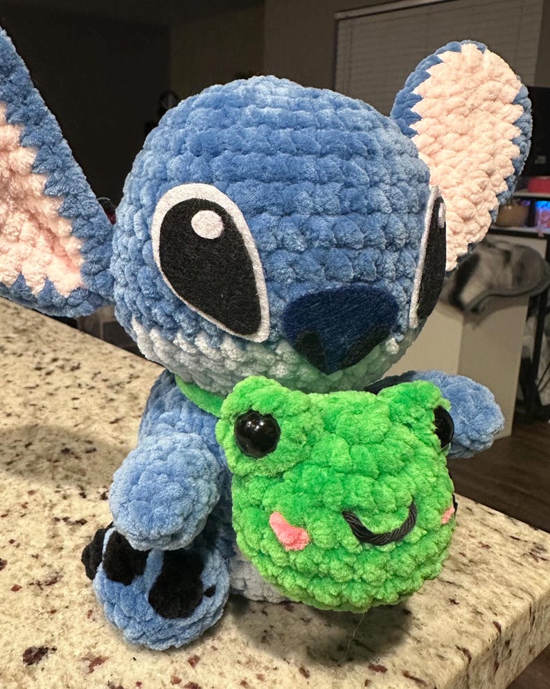 May include: A plush, blue crocheted character with large black and white eyes, pink inner ears, and a dark blue nose. It holds a green crocheted frog with black eyes and a pink mouth. The character has a green collar and black and blue feet.