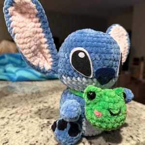May include: A blue crochet Stitch doll with large ears, black eyes, and a green collar. The doll is holding a green crochet frog with pink cheeks. The plush toy is sitting on a speckled surface.