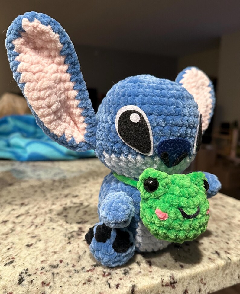 May include: A blue crochet Stitch doll with large ears, black eyes, and a green collar. The doll is holding a green crochet frog with pink cheeks. The plush toy is sitting on a speckled surface.