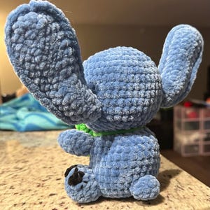 May include: A handmade blue plush toy with large ears and a green collar. The plush has a textured, velvety appearance and a small black patch on one foot. The toy is sitting on a light-colored surface.