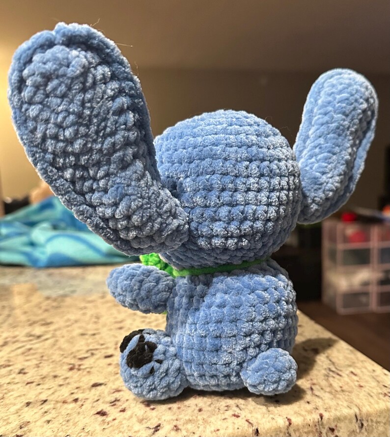 May include: A handmade blue plush toy with large ears and a green collar. The plush has a textured, velvety appearance and a small black patch on one foot. The toy is sitting on a light-colored surface.