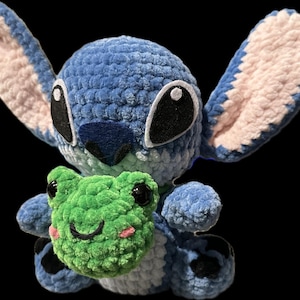 May include: A handmade blue crochet plush toy of a character with large ears, holding a green crochet frog. The plush has black eyes and a blue nose. The frog has black eyes and pink cheeks.