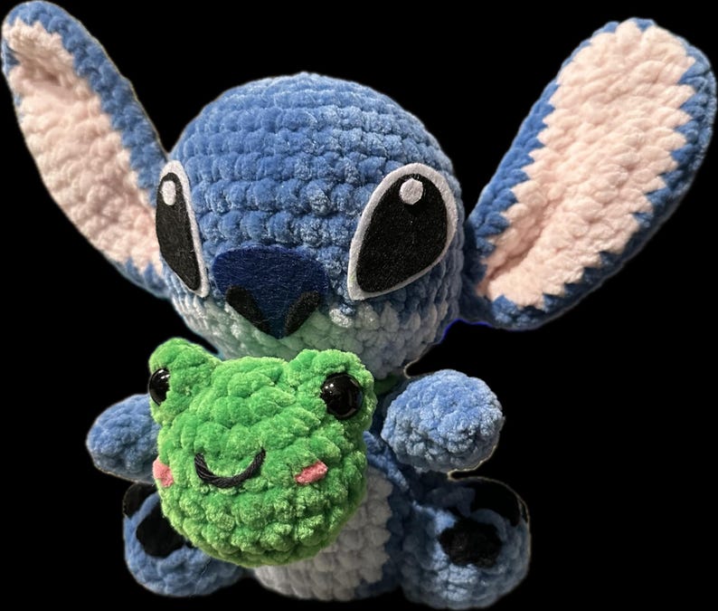 May include: A handmade blue crochet plush toy of a character with large ears, holding a green crochet frog. The plush has black eyes and a blue nose. The frog has black eyes and pink cheeks.