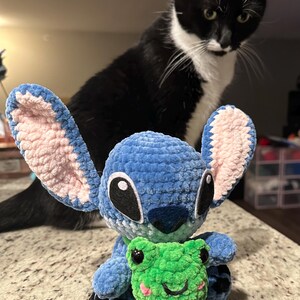 May include: A handmade blue crochet Stitch doll with pink inner ears, holding a green crochet frog. A black and white cat is in the background, looking at the doll. The doll is sitting on a speckled countertop.