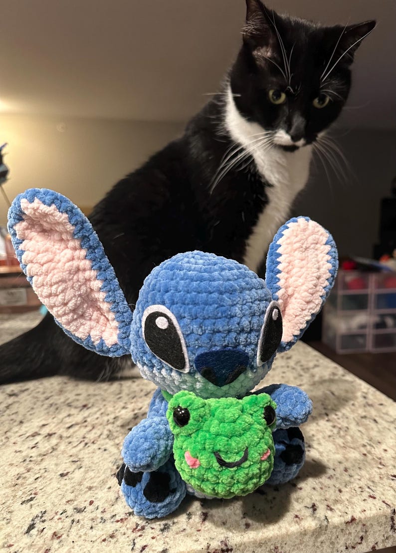 May include: A handmade blue crochet Stitch doll with pink inner ears, holding a green crochet frog. A black and white cat is in the background, looking at the doll. The doll is sitting on a speckled countertop.