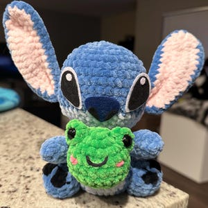 May include: A blue crocheted plush toy resembling a cartoon character with large ears, black eyes, and a dark blue nose. It is holding a green crocheted frog with a smiling face and pink cheeks. The toy is on a speckled surface.
