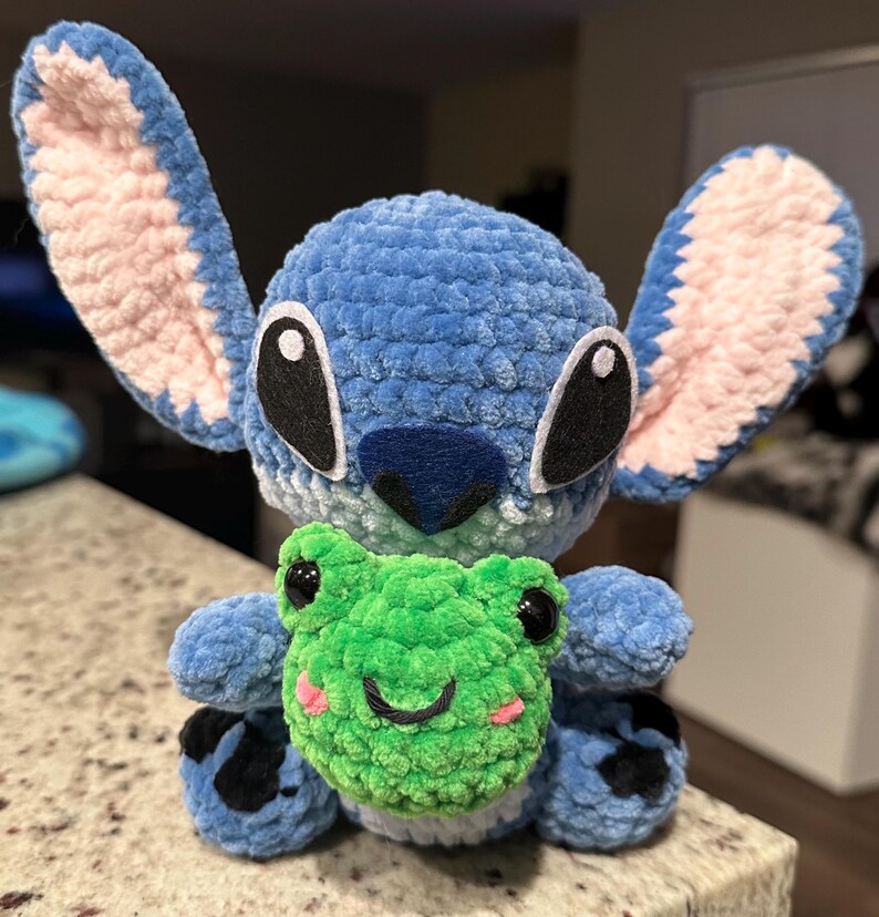 May include: A blue crocheted plush toy resembling a cartoon character with large ears, black eyes, and a dark blue nose. It is holding a green crocheted frog with a smiling face and pink cheeks. The toy is on a speckled surface.
